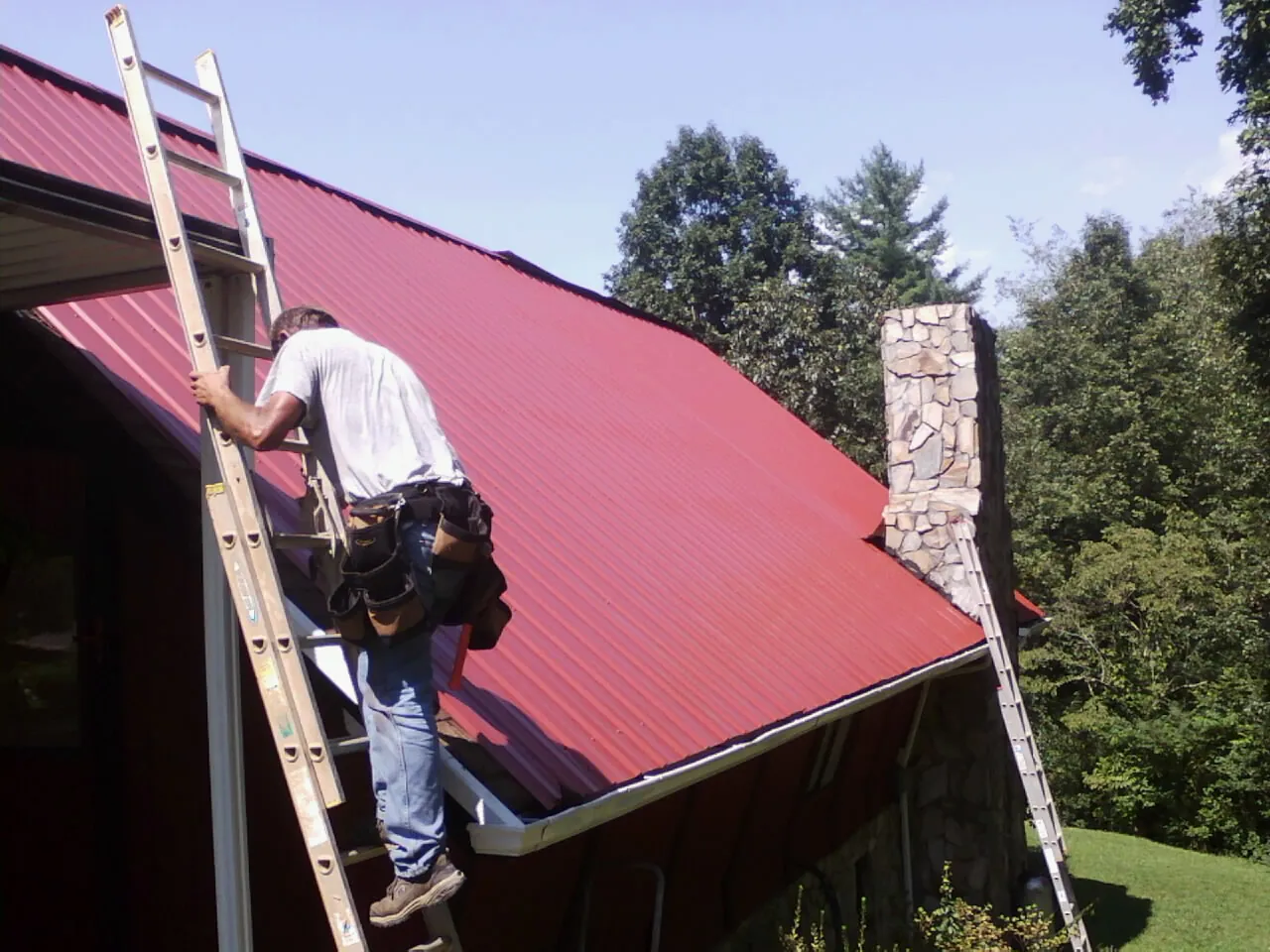 Roofer on a ladder beside a red metal roof and stone chimney in North Lewisburg